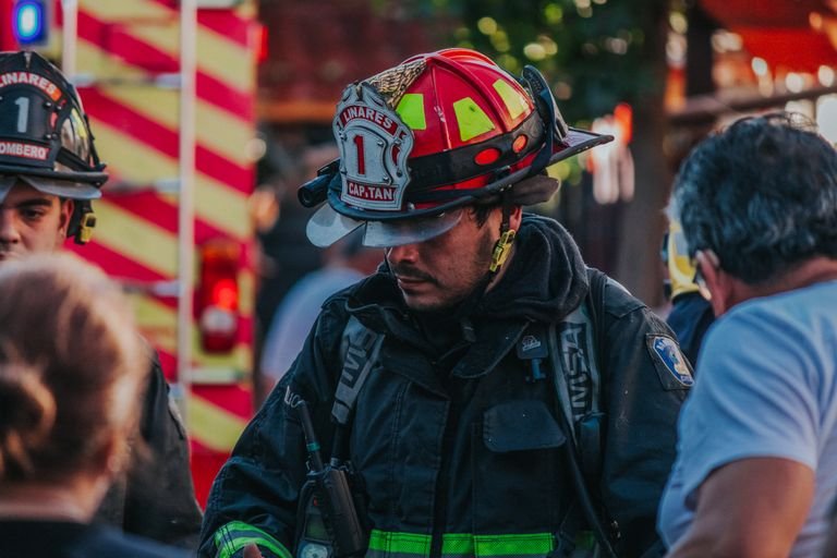 Firefighter attending an emergency on city streets, wearing uniform and helmet, with emergency vehicles in background.